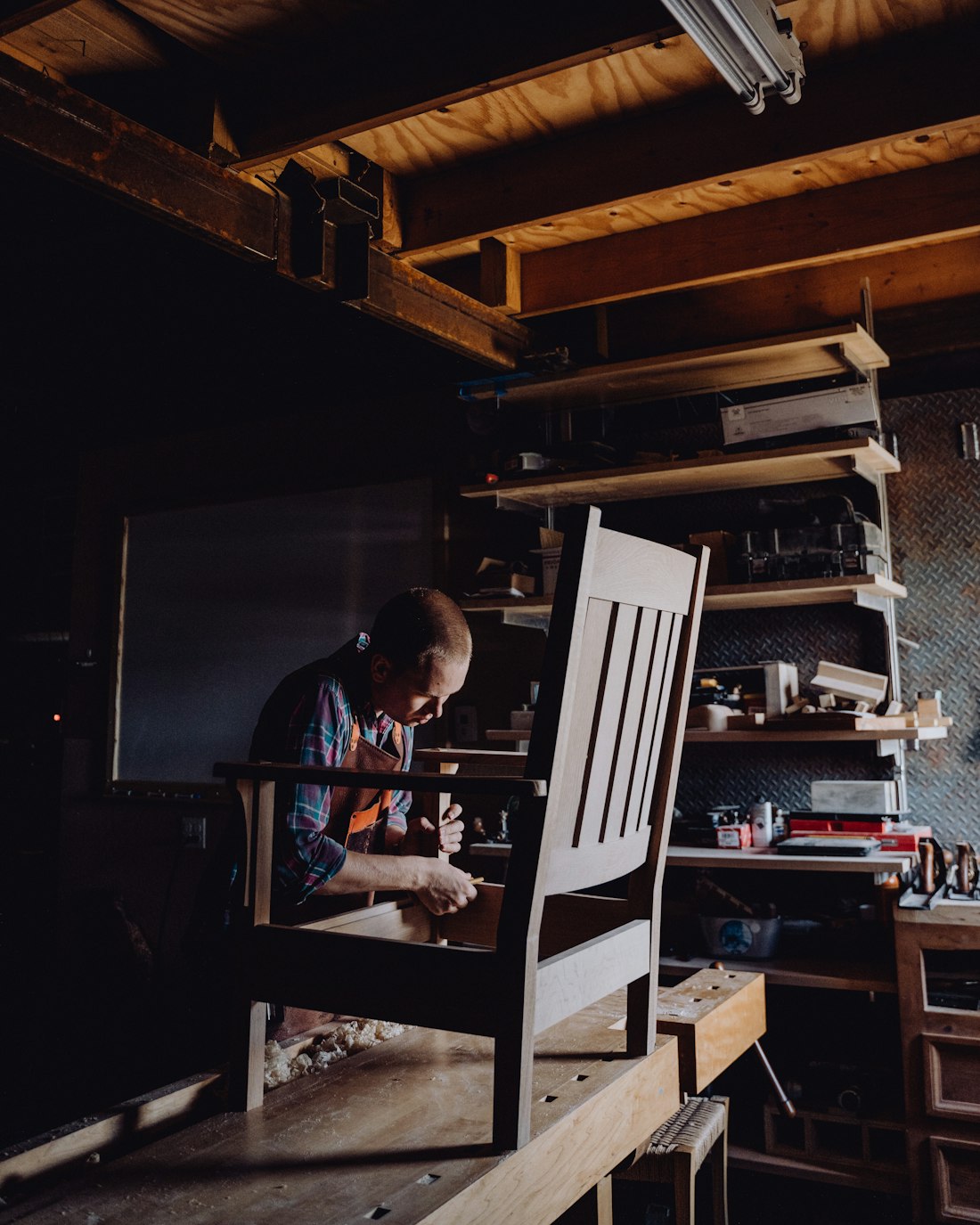 A woodworker shaping a chair part at the workbench, surrounded by tools and shavings